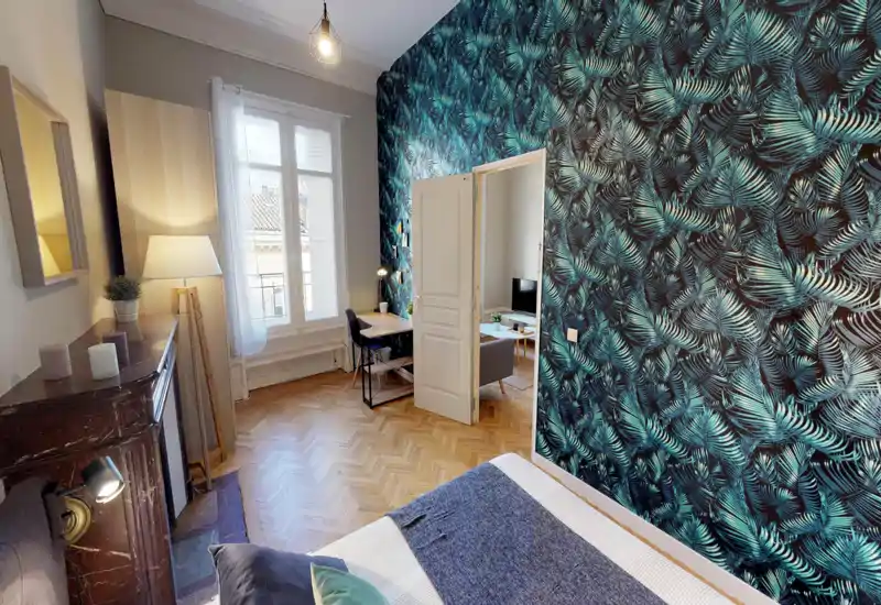 Bright, stylish bedroom with a patterned feature wall, comfortable bed dressed with cushions and a throw, and natural light from a large window. A small workspace and view into the adjacent living area make the layout functional and attractive for rentals.