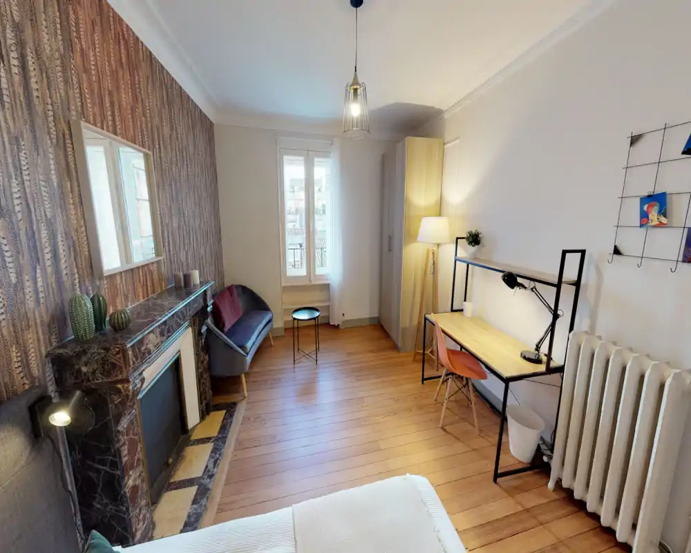 Chambre lumineuse et soignée avec parquet, papier peint décoratif et un petit coin salon. On aperçoit un coin du lit au premier plan, une armoire, un bureau et un éclairage chaud pour un confort agréable.