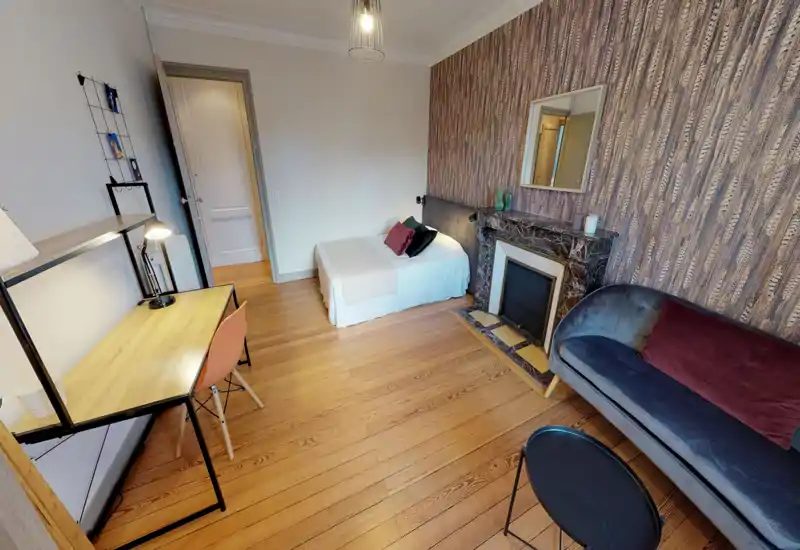 Chambre lumineuse et chaleureuse avec un lit simple, coussins décoratifs et une cheminée en marbre ; parquet et bureau pour un espace pratique.