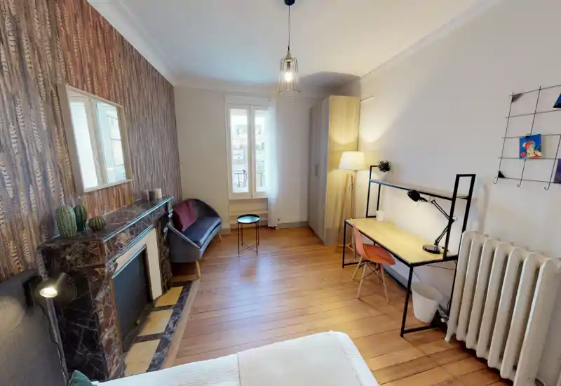 Chambre lumineuse et soignée avec parquet, papier peint décoratif et un petit coin salon. On aperçoit un coin du lit au premier plan, une armoire, un bureau et un éclairage chaud pour un confort agréable.