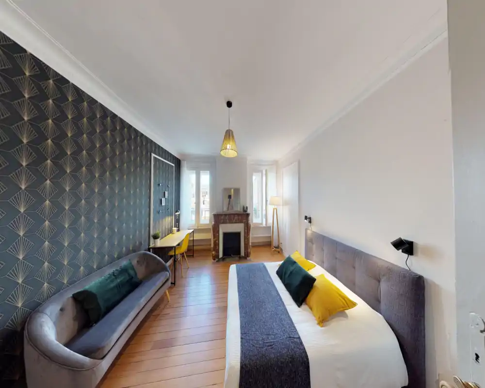 Bright, well-staged bedroom with a large double bed, decorative cushions, a patterned feature wall, a sofa and a workspace by the window — ideal for comfortable stays.