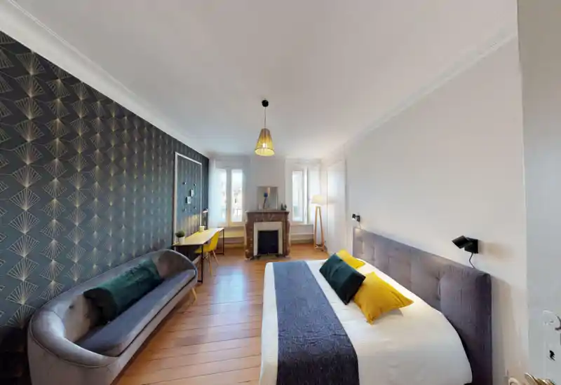 Bright, well-staged bedroom with a large double bed, decorative cushions, a patterned feature wall, a sofa and a workspace by the window — ideal for comfortable stays.