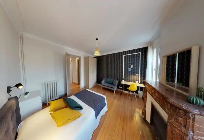Grande chambre lumineuse avec un lit double confortable, parquet chaleureux, mur à motif décoratif et un coin canapé/bureau près d'une grande fenêtre.