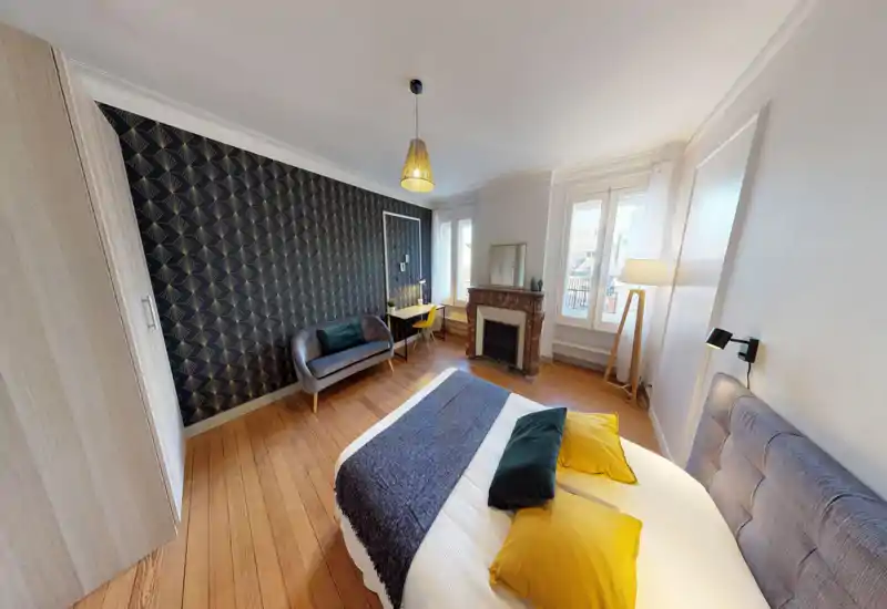 Bright, well-staged bedroom with a comfortable double bed, decorative pillows and a textured accent wall. Natural light fills the room through large windows, complemented by a cozy reading lamp and a small sofa and desk for added functionality.