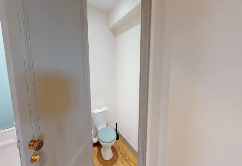 Piccolo bagno WC con vista attraverso la porta. Pareti bianche, pavimento in legno e buona luce naturale creano un ambiente semplice e ordinato.