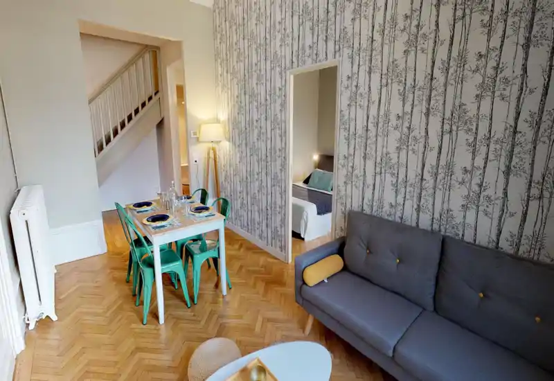 Zona giorno luminosa e di stile con carta da parati decorativa, un comodo divano grigio e un tavolo da pranzo apparecchiato—ideale per un piccolo gruppo. Pavimento in parquet e dettagli coordinati rendono l'ambiente accogliente.