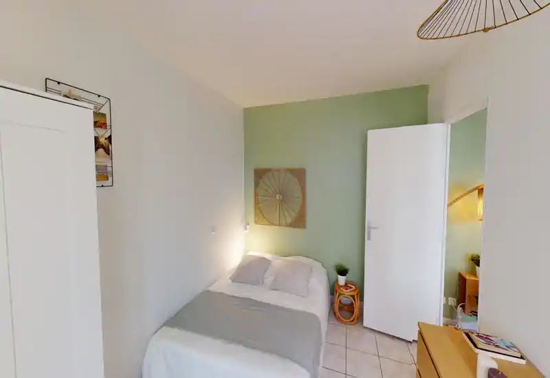 Petite chambre simple bien décorée avec lit fait, mur vert d'accent, table de chevet avec plante et décoration minimaliste. Lumineuse et accueillante — idéale pour une annonce de location.