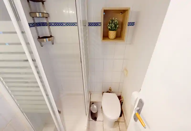 Bagno compatto e ben illuminato con doccia angolare, WC e nicchia portaoggetti; piastrelle bianche pulite e piccolo vaso decorativo.