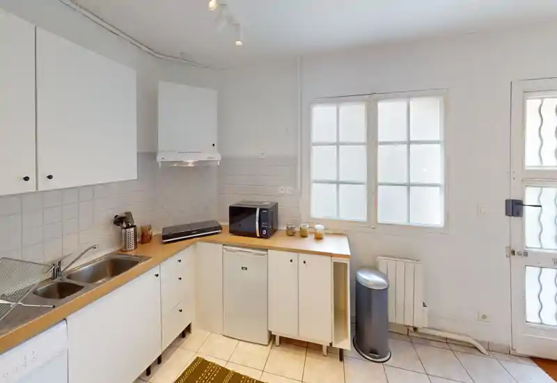 Cucina compatta e luminosa con mobili bianchi, doppio lavello, piccoli elettrodomestici sul piano e un mini-frigo — ordinata e pronta all'uso.