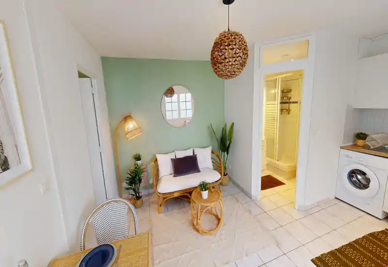Zona giorno luminosa e accogliente con poltrona in rattan, cuscini morbidi e piante decorative; layout compatto open-space con angolo cucina e porta del bagno visibili.