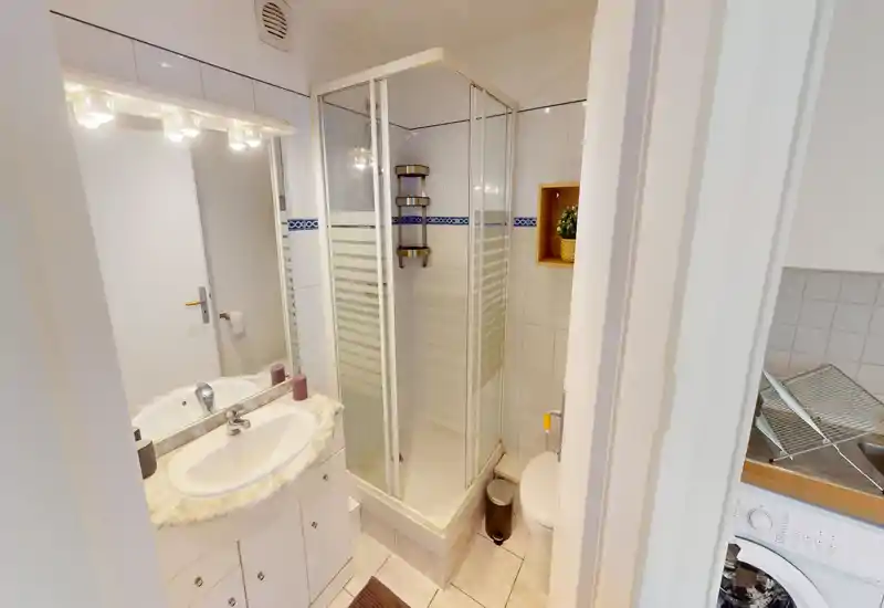 Bagno compatto e luminoso con box doccia angolare in vetro, lavabo con piano effetto marmo, specchio con luci e WC; ambiente pulito e funzionale, adatto a affitti brevi.