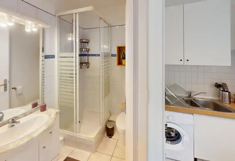 Bagno compatto e pulito con cabina doccia angolare, lavabo con specchio e mobili, e WC. L'illuminazione chiara e le piastrelle bianche donano un aspetto fresco e ordinato.