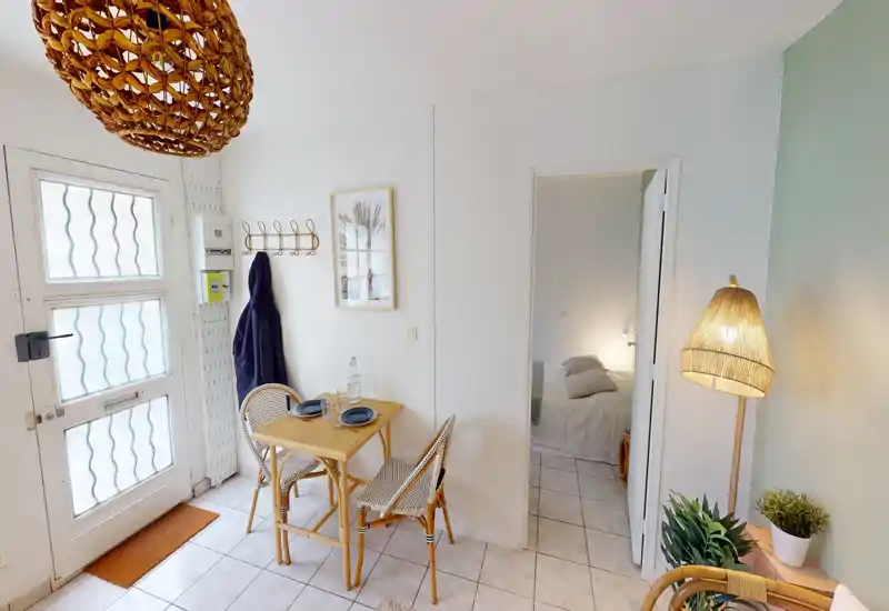 Angolo living/pranzo luminoso e accogliente con tavolo in rattan per due, lampada intrecciata e piante — ideale per un appartamento compatto.