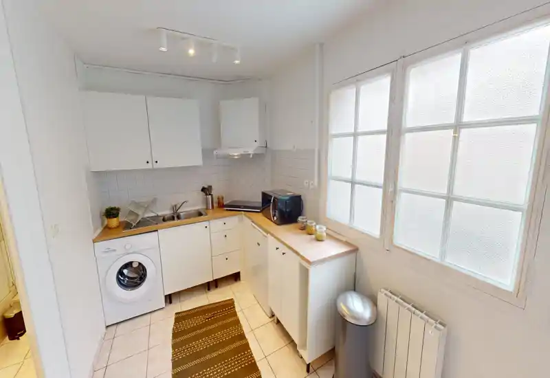Cucina compatta e ordinata con mobili bianchi, piani in legno, lavabo, lavatrice e microonde. Ampia finestra smerigliata diffonde luce naturale e un piccolo tappeto e una pianta aggiungono calore.