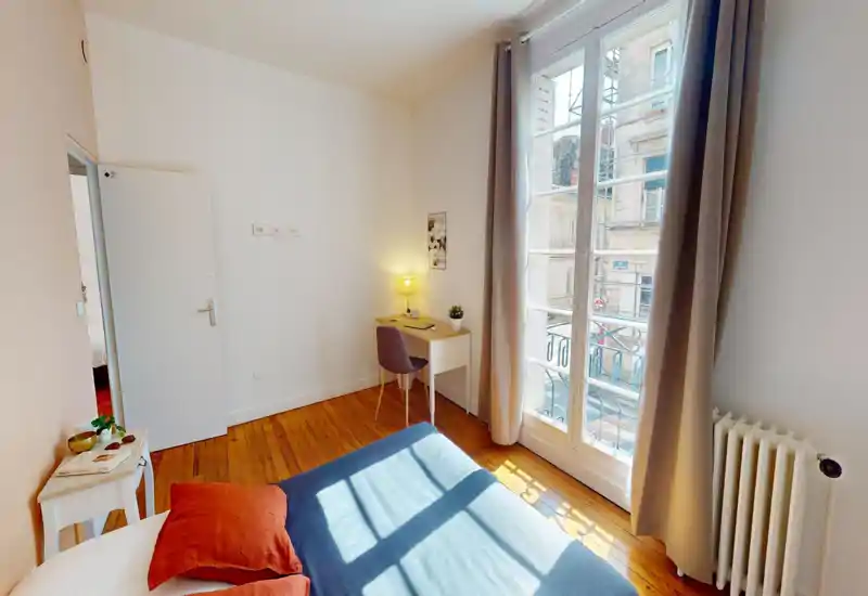 Camera da letto luminosa e accogliente con letto matrimoniale, pavimento in legno caldo e una grande finestra che lascia entrare molta luce naturale. Piccolo scrittoio con lampada accanto alla finestra per uno spazio lavoro piacevole.