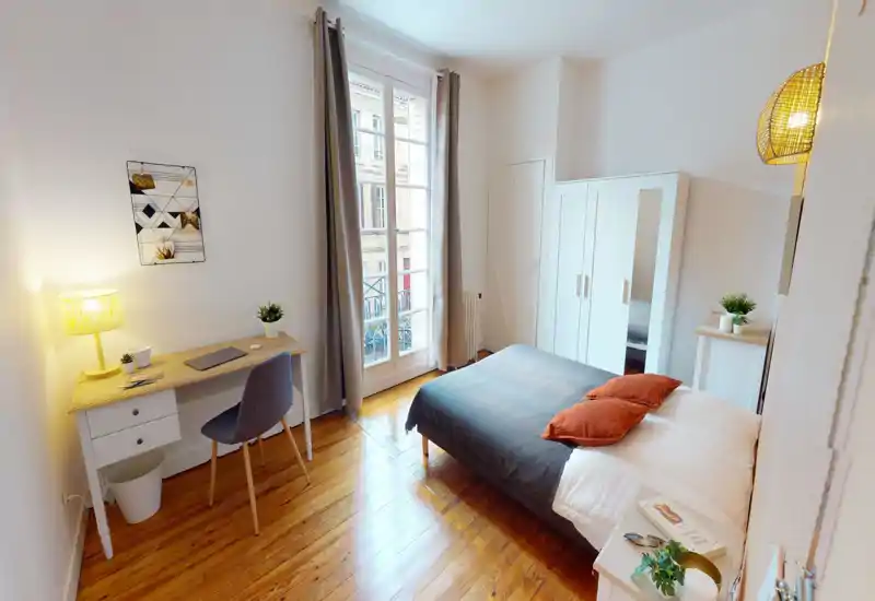 Camera da letto luminosa e ben arredata con letto matrimoniale, pavimento in legno e ampia finestra che offre molta luce naturale. La stanza dispone di scrivania, armadio e dettagli accoglienti perfetti per un soggiorno confortevole.