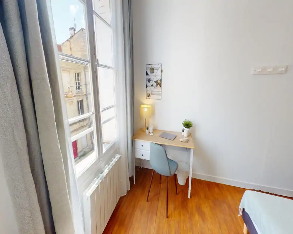 Rincón de dormitorio luminoso y sencillo con escritorio junto a una gran ventana, suelo de madera y cama parcialmente visible — acogedor y bien iluminado, ideal para una persona.