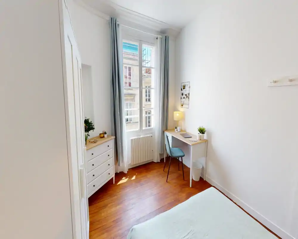 Dormitorio luminoso y ordenado con gran ventana, un escritorio pequeño y cómoda, decoración neutra y agradable — ideal para una persona o estancias cortas.