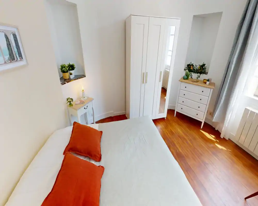 Dormitorio luminoso y acogedor con una cama amplia vestida en tonos neutros y cojines color óxido, armario blanco y cómoda junto a una ventana soleada—perfecto para estancias cómodas.