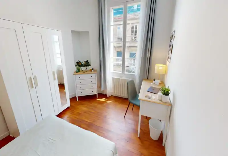 Bright, simply styled bedroom with a comfortable bed in the foreground, a wardrobe and dresser, and a desk by a large window letting in abundant natural light.