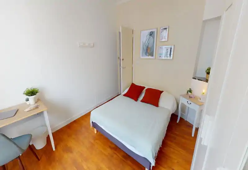 Sunlit, compact bedroom with a double bed dressed in neutral linens and two red accent pillows, a small bedside table with lamp, and a tidy desk — bright and cosy for a short stay.