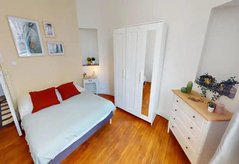 Bright, cozy bedroom with a double bed dressed in neutral linens, two rust-coloured cushions, a white wardrobe with mirror, and a chest of drawers topped with plants and decor.
