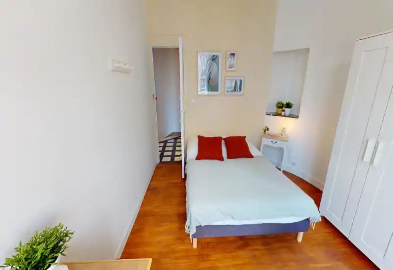 Bright and tidy bedroom with a double bed, warm wooden floor and simple, modern decor. Two red accent pillows and a bedside table with plants make the room inviting — ideal for a comfortable short stay.