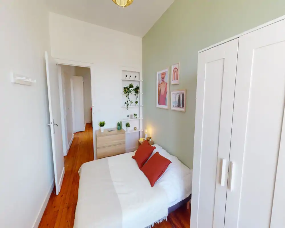 Sunny, well-styled bedroom with a double bed, warm wooden floor, white wardrobe and soft green accent wall decorated with framed prints and plants.