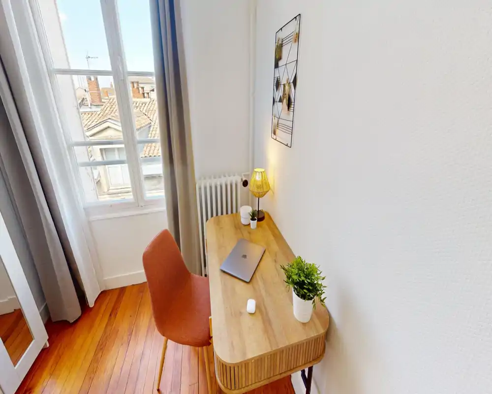 Bright corner living/working area with a wooden desk, cozy chair, lamp and plants by a large window providing natural light — a calm spot for remote work or reading.