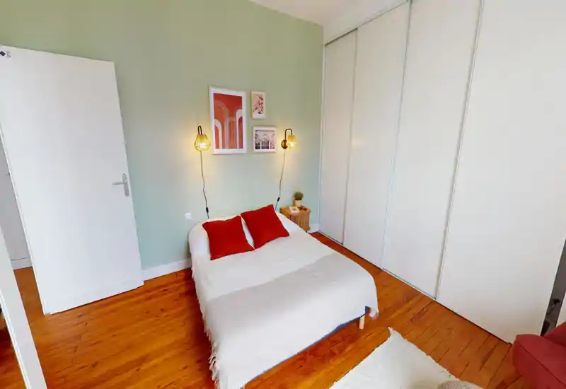 Camera da letto accogliente e luminosa con letto matrimoniale ben rifatto, coperta morbida e due cuscini rossi, quadri decorativi e armadi scorrevoli bianchi incassati.