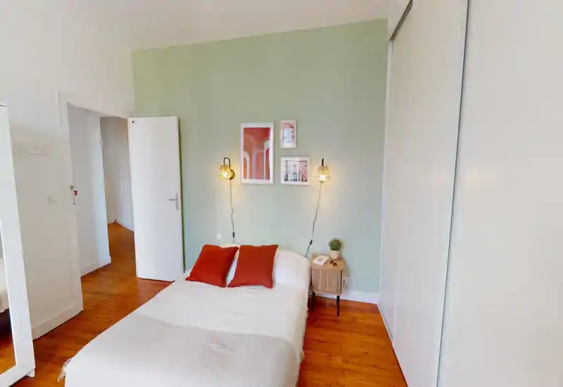 Camera da letto luminosa e curata con letto matrimoniale centrale, pavimento in legno caldo e luci da parete soffuse. Cuscini rossi e quadri rendono l’ambiente accogliente e moderno.