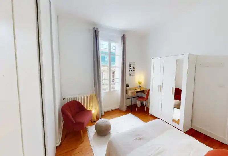 Camera da letto luminosa e accogliente con finestra grande, biancheria neutra, armadio bianco con specchio, scrivania e poltrona rossa: ideale per soggiorni confortevoli e smart working.