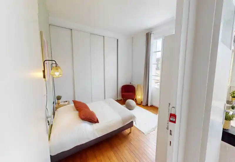 Camera da letto luminosa e accogliente con letto matrimoniale in lino bianco e cuscini terracotta, lampada da comodino, armadi a muro e poltrona vicino alla finestra.