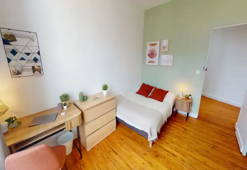 Camera da letto luminosa e curata con un comodo letto matrimoniale in lenzuola neutre, due cuscini color ruggine, un comodino con lampada, scrivania e cassettiera in legno — pavimento caldo e parete accentata verde chiaro.