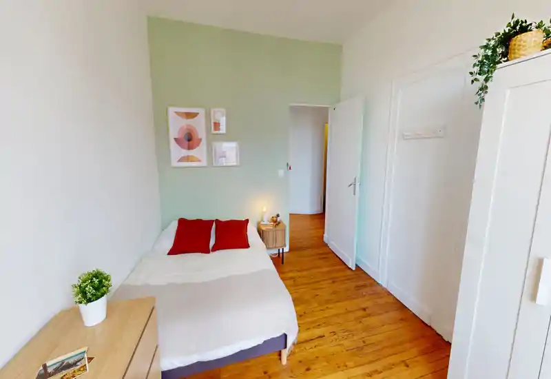 Camera da letto luminosa e ben arredata con letto matrimoniale in lenzuola neutre e due cuscini rossi, pavimento in legno e quadri minimal alle pareti. Un comodino con lampada e un armadio offrono spazio e funzionalità in un ambiente arioso.