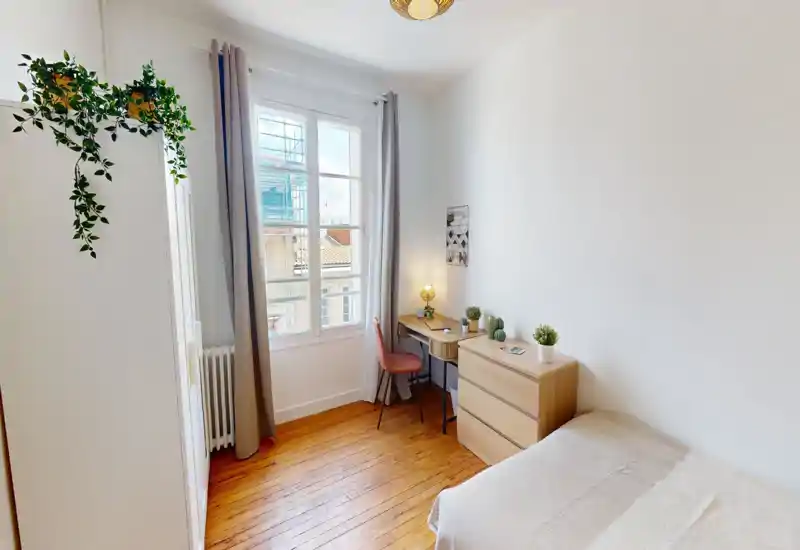 Camera da letto singola luminosa e ordinata con letto nella parte inferiore destra, pavimenti in legno, scrivania e comò accanto a una grande finestra che illumina la stanza.