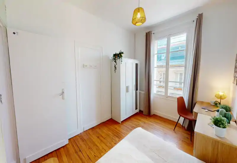 Camera da letto luminosa e ben arredata con letto visibile in primo piano, pavimenti in legno, armadio bianco e scrivania vicino a una grande finestra che illumina la stanza.