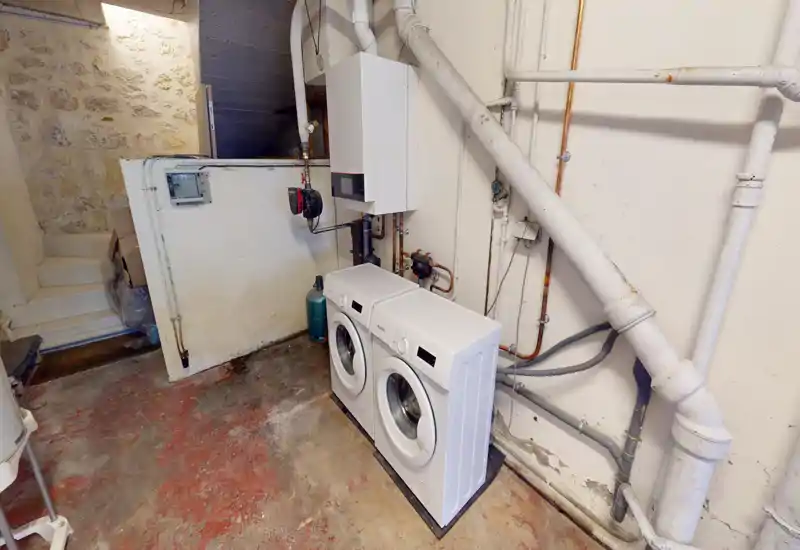 Locale tecnico/lavanderia con due lavatrici a carica frontale, caldaia a parete e tubazioni a vista. Spazio funzionale con pavimento in cemento, ideale per uso di servizio più che abitativo.