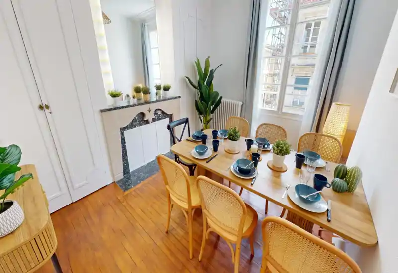 Sala da pranzo luminosa e stilosa con tavolo in legno apparecchiato, sedie in midollino e una grande finestra che offre molta luce naturale.