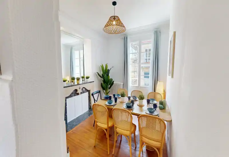Sala da pranzo luminosa e curata con tavolo in legno per sei persone, sedie in midollino e piante decorative. Ampia finestra con tende chiare che garantisce molta luce naturale e un'atmosfera accogliente.