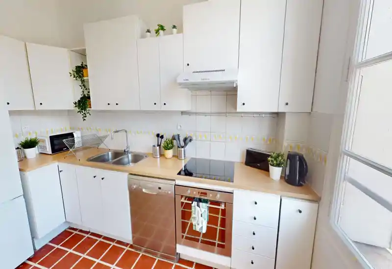 Cucina luminosa e ordinata con pensili bianchi, forno e lavastoviglie incassati, doppio lavello e piccole piante decorative. Ampia finestra che dona luce naturale e piano di lavoro ben organizzato.