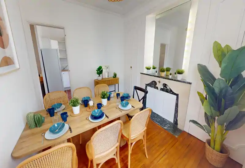 Sala da pranzo luminosa e curata con tavolo in legno apparecchiato per più persone, sedie in giunco e piante decorative per un'atmosfera accogliente.