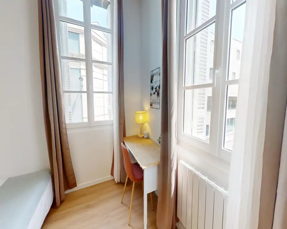 Angolo di camera luminoso e compatto con ampie finestre, una piccola scrivania e area notte. Il bordo del materasso è visibile a sinistra, mentre una lampada calda e la sedia creano un angolo lavoro accogliente.