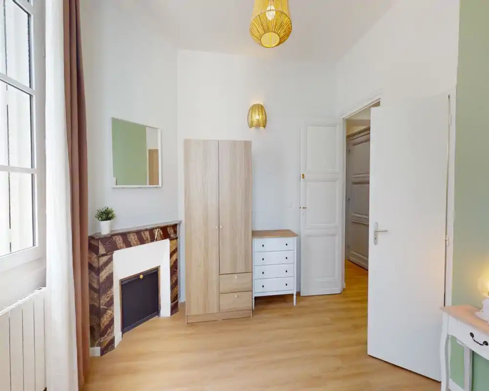 Angolo di camera luminoso e minimal con pavimento in legno, armadio e comò, applique decorativa e un piccolo camino in marmo accanto a una grande finestra che illumina la stanza.