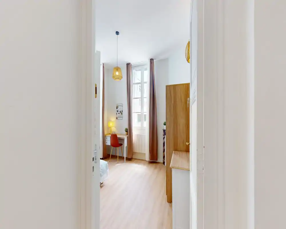 Camera da letto luminosa e moderna con luce naturale, una scrivania e una sedia vicino alla finestra, lampade sospese calde e letto parzialmente visibile — perfetta come singola o stanza per ospiti.