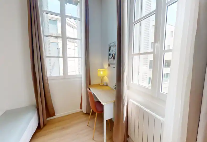 Angolo di camera luminoso e compatto con ampie finestre, una piccola scrivania e area notte. Il bordo del materasso è visibile a sinistra, mentre una lampada calda e la sedia creano un angolo lavoro accogliente.