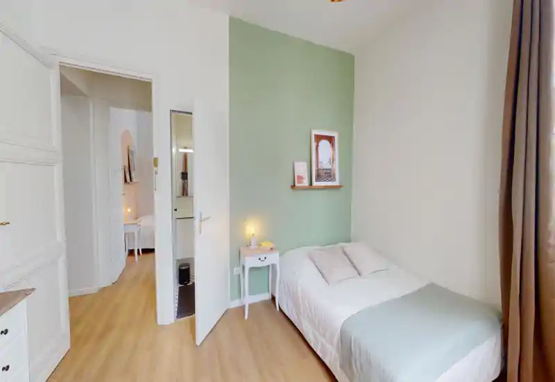 Camera da letto luminosa e ordinata con letto singolo contro una parete verde chiaro, comodino con lampada e stampe incorniciate — perfetta per un soggiorno accogliente.