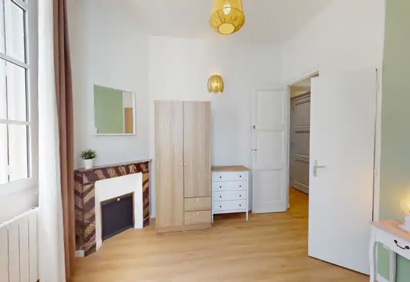 Coin de chambre lumineux et sobrement meublé avec armoire, commode, cheminée décorative et grande fenêtre laissant entrer une belle lumière naturelle.