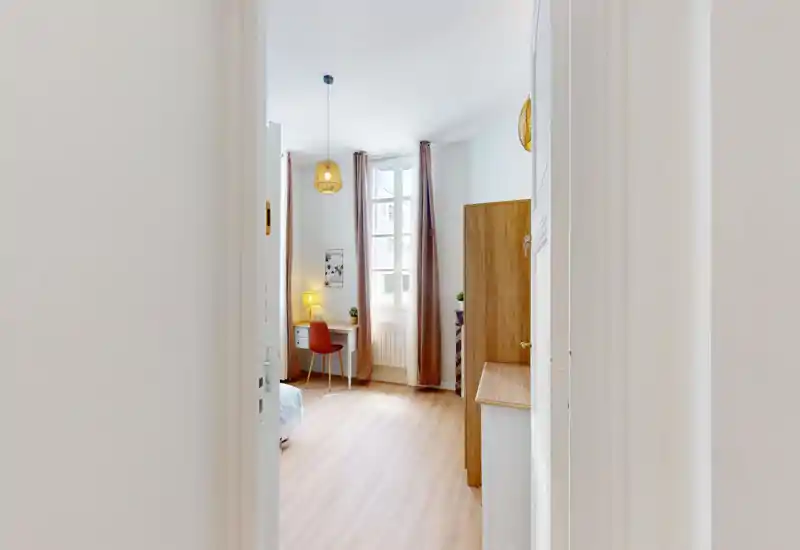 Camera da letto luminosa e moderna con luce naturale, una scrivania e una sedia vicino alla finestra, lampade sospese calde e letto parzialmente visibile — perfetta come singola o stanza per ospiti.