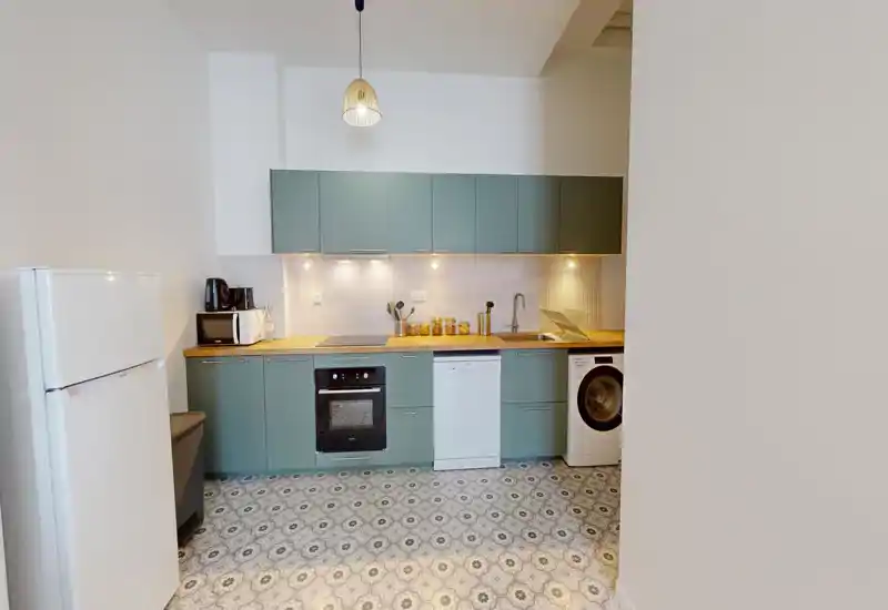 Cucina moderna e compatta con mobili verdi, top in legno, forno integrato, lavastoviglie e lavatrice; luce luminosa e sospensione calda che aggiunge un tocco accogliente.